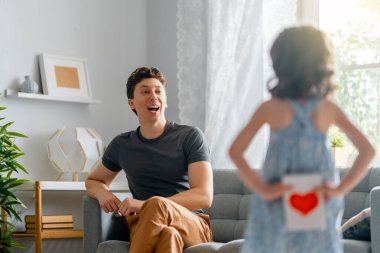 Çocuk kızı babasını tebrik ediyor ve ona kartpostal veriyor. Baba ve kız gülümsüyor. Aile tatili ve birliktelik.