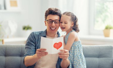 Çocuk kızı babasını tebrik ediyor ve ona kartpostal veriyor. Baba ve kız gülümsüyor ve sarılıyorlar. Aile tatili ve birliktelik.