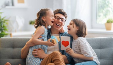 Çocuklar babalarını tebrik ediyor ve ona kartpostal veriyorlar. Baba ve kızlar gülümsüyor ve sarılıyorlar. Aile tatili ve birliktelik.