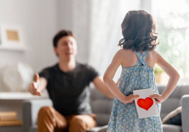 Çocuk kızı babasını tebrik ediyor ve ona kartpostal veriyor. Baba ve kız gülümsüyor. Aile tatili ve birliktelik.