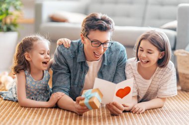 Çocuklar babalarını tebrik ediyor ve ona kartpostal veriyorlar. Baba ve kızlar gülümsüyor ve sarılıyorlar. Aile tatili ve birliktelik.