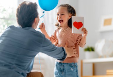 Çocuk kızı babasını tebrik ediyor ve ona kartpostal veriyor. Baba ve kız gülümsüyor ve sarılıyorlar. Aile tatili ve birliktelik.