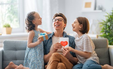 Çocuklar babalarını tebrik ediyor ve ona kartpostal veriyorlar. Baba ve kızlar gülümsüyor ve sarılıyorlar. Aile tatili ve birliktelik.