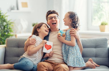 Çocuklar babalarını tebrik ediyor ve ona kartpostal veriyorlar. Baba ve kızlar gülümsüyor ve sarılıyorlar. Aile tatili ve birliktelik.