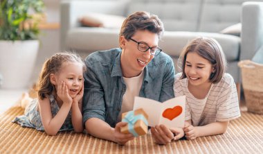 Çocuklar babalarını tebrik ediyor ve ona kartpostal veriyorlar. Baba ve kızlar gülümsüyor ve sarılıyorlar. Aile tatili ve birliktelik.