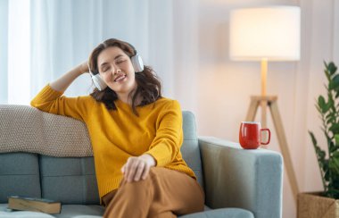 Neşeli genç bir kadın kulaklıkla müzik dinliyor evdeki kanepede oturuyor..