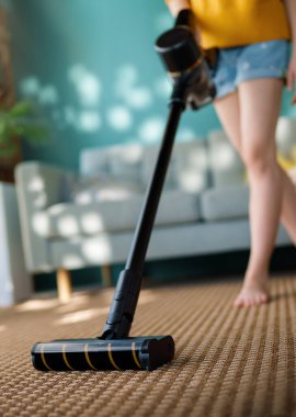 Oturma odasında kablosuz elektrik süpürgesiyle yerleri süpüren genç bir kadının fotoğrafını çek. Ev ve kullanışlı teknoloji kavramı.