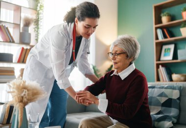 Doktor yaşlı kadının ayağa kalkmasına yardım ediyor..