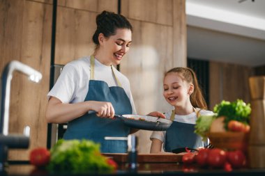 Modern bir mutfakta sağlıklı bir yemek pişirirken neşeli anne-kız kaynaşıyor. Her ikisi de uyumlu mavi önlükler giyer ve etrafları taze sebzelerle çevrilidir. Aile birlikteliğini, sağlıklı yaşamı ve aşçılık yaratıcılığını kutlarlar..