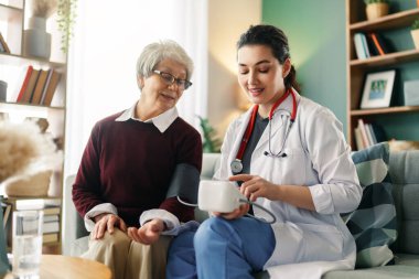 İlgili bir hemşire, rahat bir evde yaşlı bir kadının tansiyonunu ölçmesine yardımcı olur. Bu resim profesyonel sağlık hizmetlerini, hasta bakımını ve düzenli sağlık kontrollerinin önemini vurgulayarak, sıcak ve güven verici bir atmosfer sunmaktadır..