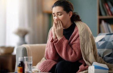 Battaniyeye sarılı bir kadın yüzüne mendil tutarak rahat bir kanepeye oturur. Çevreler arasında ilaçlar ve dokular, soğuk algınlığı sırasında kendine bakmanın vurgulanması yer alıyor. Sıcak ortam rahatlatıcı bir ev ortamı sunuyor..