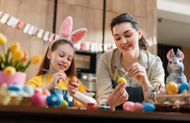 Neşeli anne ve kız Paskalya yumurtalarını parlak bir mutfakta boyayarak güzel vakit geçiriyorlar. Küçük kız neşeli tavşan kulakları takıyor ve neşeli, neşeli bir tatil ortamı yaratıyor. Canlı bahar laleleri ve dekoratif yumurtalarla çevrili..
