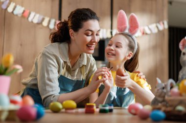 Neşeli anne ve kızı neşeli bir mutfakta renkli Paskalya yumurtaları boyarken kaynaşıyorlar. Küçük kız eğlenceli tavşan kulakları takıyor ve ikisi de yaratıcı, neşeli bir anın tadını çıkarıyor. Aile geleneklerinin mükemmel bir temsili..