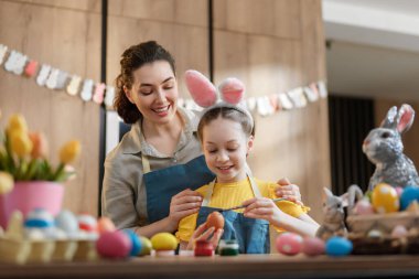 Neşeli bir anne ve kızı, tavşan kulağı takmış, sıcacık bir mutfakta Paskalya yumurtalarını birlikte boyuyorlar. Renkli yumurtalar ve bahar süslemeleriyle çevrelenmiş kahkahaları ve bağlantıları ailenin ruhunu somutlaştırıyor..
