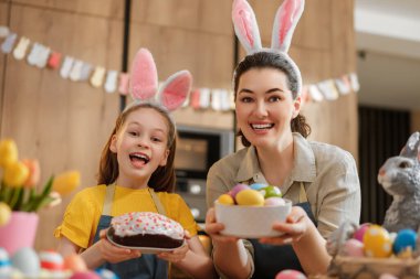 Tavşan kulağı takan neşeli anne ve kız birlikte Paskalya 'yı kutluyor, bir kase renkli yumurta ve ev yapımı kek tutarken gülüşmeler ve kahkahalar atıyorlar. Parlak dekorasyonlar ve taze laleler sıcak, şenlikli bir atmosfer yaratır..
