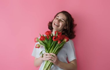 Canlı pembe bir arka planda duran canlı bir buket kırmızı lale tutan bu genç bayanın sevincinin sevinci. Işıl ışıl gülüşü ve rahat duruşu, baharı kutlamak için mükemmel bir mutluluk ve tazelik hissi veriyor..