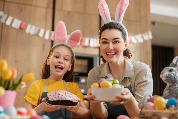 Tavşan kulağı takan neşeli anne ve kız birlikte Paskalya 'yı kutluyor, bir kase renkli yumurta ve ev yapımı kek tutarken gülüşmeler ve kahkahalar atıyorlar. Parlak dekorasyonlar ve taze laleler sıcak, şenlikli bir atmosfer yaratır..