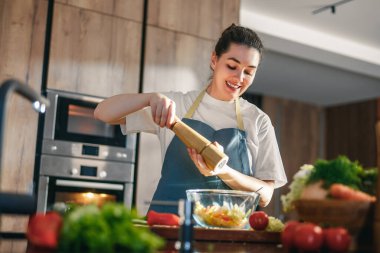 Önlüklü neşeli bir kadın, çağdaş bir mutfakta taze bir salataya baharat katar. Sahne, taze malzemeler ve sıcak bir atmosferle yemek pişirmenin ve sağlıklı yaşamanın neşesini yakalıyor..