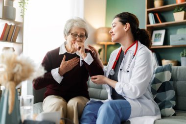 Sıcak, davetkar bir oturma odasında yaşlı bir kadının sağlığıyla ilgilenen şefkatli bir kadın doktor. Sahne, evde tedavi görmenin, empati göstermenin, destek vermenin ve bir sağlık uzmanı arasındaki kişisel bağın özünü yakalıyor.