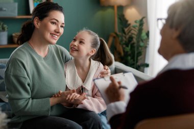Bir anne ve kızı terapistle terapi seansına katılıyor. Ortam sakin ve davetkar, duygusal destek ve iyileşme vurgulanıyor..