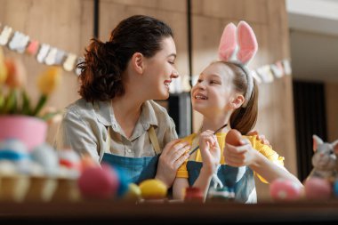 Neşeli anne ve kızı Paskalya yumurtalarını parlak bir mutfakta boyarken birbirlerine bağlanıyorlar. Önlükler ve tavşan kulakları takarak gülümsüyorlar ve neşeli dekorasyonlarla çevrili yaratıcı aile zamanlarından zevk alıyorlar. Birliktelik teması için mükemmel, ünlüler.