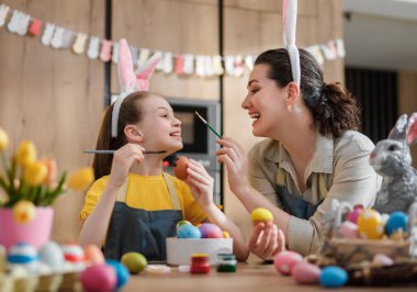 Neşeli bir anne ve kız, tavşan kulağı takıyor, sıcak bir mutfak ortamında paskalya yumurtalarını neşeyle boyuyor. Sahne, Paskalya bayramında aile bağlarının neşe ve yaratıcılığını temsil eden canlı renklerle dolu. İdeal