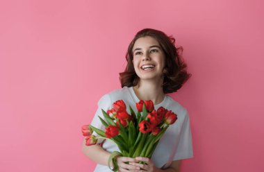 Neşeli genç bir kadın elinde canlı bir buket kırmızı lale tutarken neşeyle güler. Parlak pembe bir zemin, gülüşü ve rahat duruşu mutluluk ve pozitiflik saçıyor. Bahar temalı promosyonlar, tebrik kartları ya da yaşam tarzı için mükemmel.