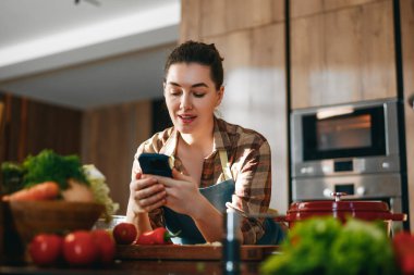 Modern bir mutfakta akıllı telefon kullanan ve taze sebze hazırlayan neşeli bir kadın. Sıcak ışıklandırma ve ahşap dolap rahat, davetkar bir atmosfer yaratıyor, yemek blogları ve yemek pişirme içeriği için mükemmel..