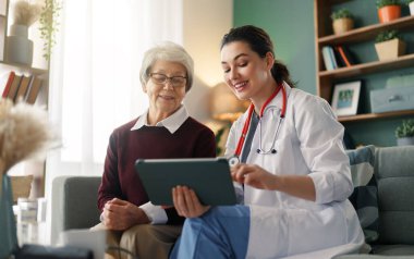 A compassionate female doctor in a comforting home environment shares digital healthcare information with a smiling elderly woman. The scene depicts empathy, advanced healthcare, and interpersonal connection, highlighting the importance of technology