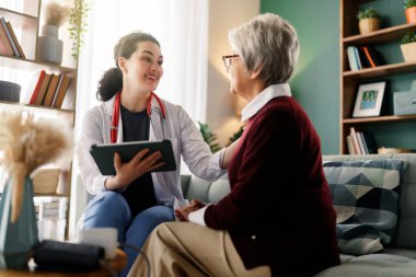 A compassionate doctor with a stethoscope and digital tablet engages with an elderly patient during a home visit. The warm, modern setting fosters an environment of trust and personalized healthcare.