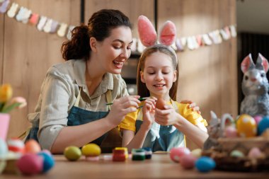 Sıcak, sade bir mutfak ortamında paskalya yumurtası resmiyle mutlu bir anne-kız ilişkisi. Çocuk şirin tavşan kulakları takıyor ve neşeli atmosfere neşe katıyor. Renkli yumurtalar ve bahar süslemeleriyle çevrili, bu iç açıcı anne