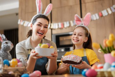 Neşeli bir anne ve kız birlikte Paskalyayı kutluyor, tavşan kulağı takıyor ve parlak bir mutfakta renkli yumurtalar ve ev yapımı pasta sergiliyor. Atmosfer neşe, aile bağları ve şenlik ruhuyla dolu..