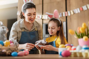 Neşeli anne ve kız tavşan kulağı ve önlük takıyor, parlak bir mutfakta birlikte pasta süslüyorlar. Renkli Paskalya yumurtaları, çiçekler ve şenlikli dekorlarla çevrelenmiş bu iç açıcı görüntü ailenin ruhunu, kutlamasını ve baharını yansıtıyor.