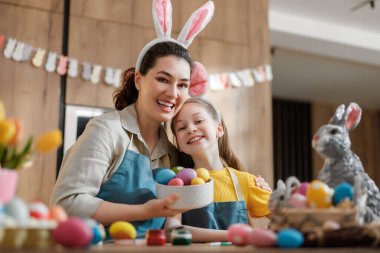 Joyful mother and daughter wearing bunny ears and aprons, smiling as they prepare vibrant Easter eggs together in a cozy kitchen. This heartwarming image captures genuine happiness, family bonding, and creative holiday traditions, perfect for promoti