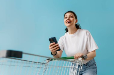 Günlük beyaz tişörtlü neşeli genç bir kadın akıllı telefonunu bir alışveriş arabasını iterken kullanıyor, canlı mavi bir arka plana pozitif enerji ve heyecan yayıyor. Reklamlar, perakende reklamlar, market teslimatları, alışveriş uygulamaları için mükemmel.