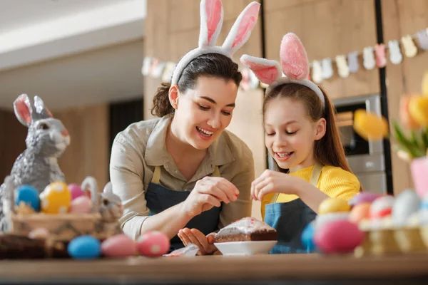 Neşeli bir anne-kız takımı Paskalya Bayramı 'nda bir pasta süsledi. Her ikisi de renkli Paskalya yumurtaları ve bahar süsleriyle çevrili neşeli tavşan kulakları takıyor. Bu iç açıcı sahne aile bağlarının ve yaratıcılığın özünü yakalıyor.