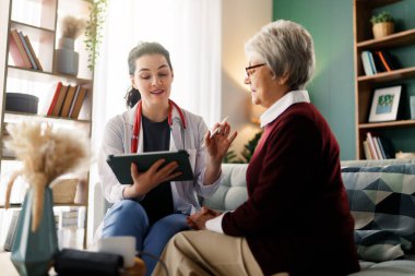 Şefkatli genç bir doktor ev ziyareti sırasında yaşlı bir hastaya danışıyor. Ortam sıcak ve davetkar, sağlık hizmetlerindeki güven ve ilgiyi sembolize ediyor. Tıbbi danışma, yaşlı bakımı ve ev sağlığı hizmetleri konuları için mükemmel.