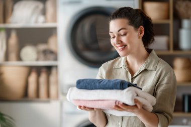 Neşeli genç bir kadın, temiz ve katlanmış çamaşırların taze kokusunun ve rahatlatıcı hissinin tadını çıkarıyor. Etrafı güneş ışığıyla çevrili, memnun ve memnun görünüyor, evde bakımı, temizliği ve dengeli bir hayatı simgeliyor.