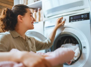 Neşeli kadın, aydınlık ve düzenli bir çamaşırhanede çamaşır yıkamayı seviyor. Modern bir çamaşır makinesini çalıştırırken gülümsüyor, rahat bir ev ortamında başarı, günlük rutin ve rahatlık hissi veriyor. Bu ticari stok fotoğrafı.