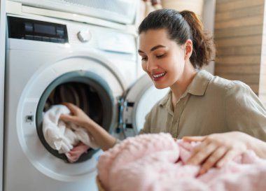Neşeli genç bir kadın ev hayatının tadını çıkarırken giysileri modern bir çamaşır makinesine yüklüyor. Parlak ve rahat çamaşır odası atmosferi günlük rutini tatmin, rahatlık ve temizlikle vurguluyor. Mükemmel.
