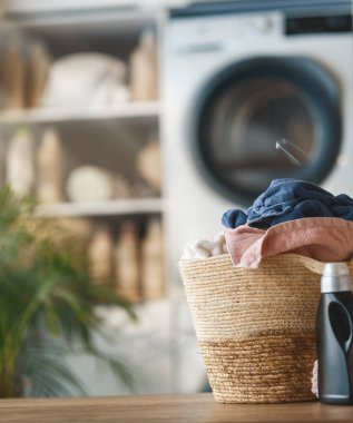 Sıcak, davetkar çamaşır odası sahnesinde yeni yıkanmış havlu ve kıyafetlerle dolu şık bir hasır sepet var. Arka plandaki modern çamaşır makinesi temiz ve düzenli ev ortamını geliştirir. Güneşli iç mekan, doğal dokular ve ev.