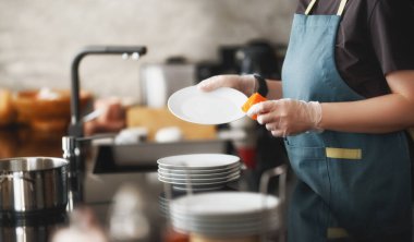 Çağdaş bir mutfakta tabak fırçalarken eldiven ve önlük giyen birinin yakın çekimi. Görüntü temizliği, ev bakımını ve hijyene olan ilgiyi aktarıyor. Ev işleri, ticari temizlik hizmetleri, sağlık hizmetleri için mükemmel.