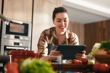 Genç bir kadın, aydınlık ve modern bir mutfakta yemek pişirirken neşeyle tablet kullanıyor. Taze sebzeler ve rahat bir ortam ile çevrelenmiş halde, teknoloji ve mutfak sanatlarının birleşimini kucaklıyor..