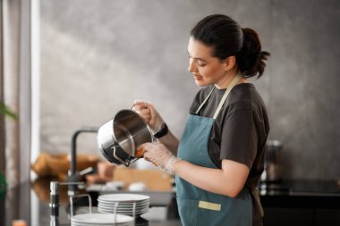 Gündelik kıyafetler ve önlükle neşeli bir kadın modern bir mutfakta paslanmaz çelik bir tencereyi yıkıyor. Sahne temizlik, özen ve günlük rutin ışık saçıyor. Gülüşü ve rahat duruşu, ho 'da memnuniyet, sorumluluk ve gurur hissi uyandırıyor.