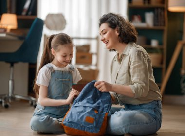 Bir anne ve kızın mavi bir sırt çantasını güzel aydınlatılmış, rahat bir ev ortamında yere oturttuğu neşeli bir aile bağı anı. Bu iç açıcı sahne ebeveyn bakımını, çocukluk mutluluğunu ve günlük aile rutinlerini yansıtıyor, Evo.