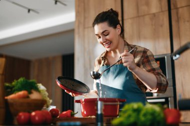 Modern bir mutfakta yemek pişirmekten zevk alan neşeli bir kadın. Taze sebzelerle çevrelenmiş halde, yemek hazırlamak için kırmızı bir tencere ve kepçe kullanıyor, ev yemeği pişirme sanatını ve mutfak yaratıcılığını örnek alıyor..