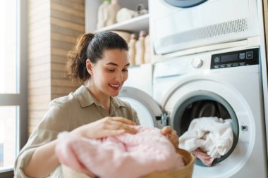 Modern bir çamaşır odasında çamaşır makinesinden taze, yumuşak çamaşırları çıkarırken ev işlerinin keyfini çıkaran neşeli genç bir kadın..