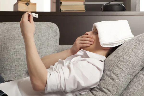 Kranker junger Mann liegt auf Sofa und überprüft seine Temperatur — Stockfoto