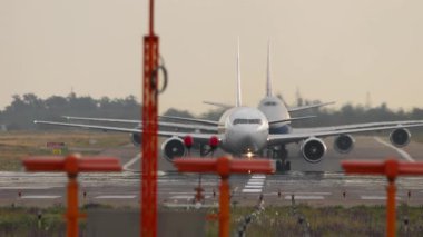 İki ticari uçak pistte sırayla hareket ediyor, ön taraftan görülüyor. Uçaklar yavaş hareket ediyor, kalkışa hazırlanıyorlar. Hava yolculuğu temaları için uygun