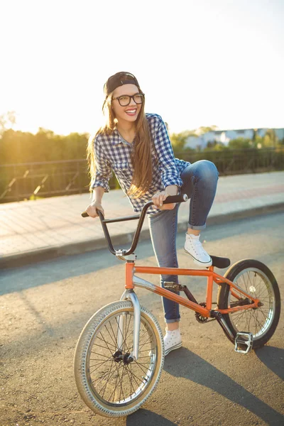 Bella giovane donna in un cappello in sella a una bicicletta su sfondo città — Foto Stock